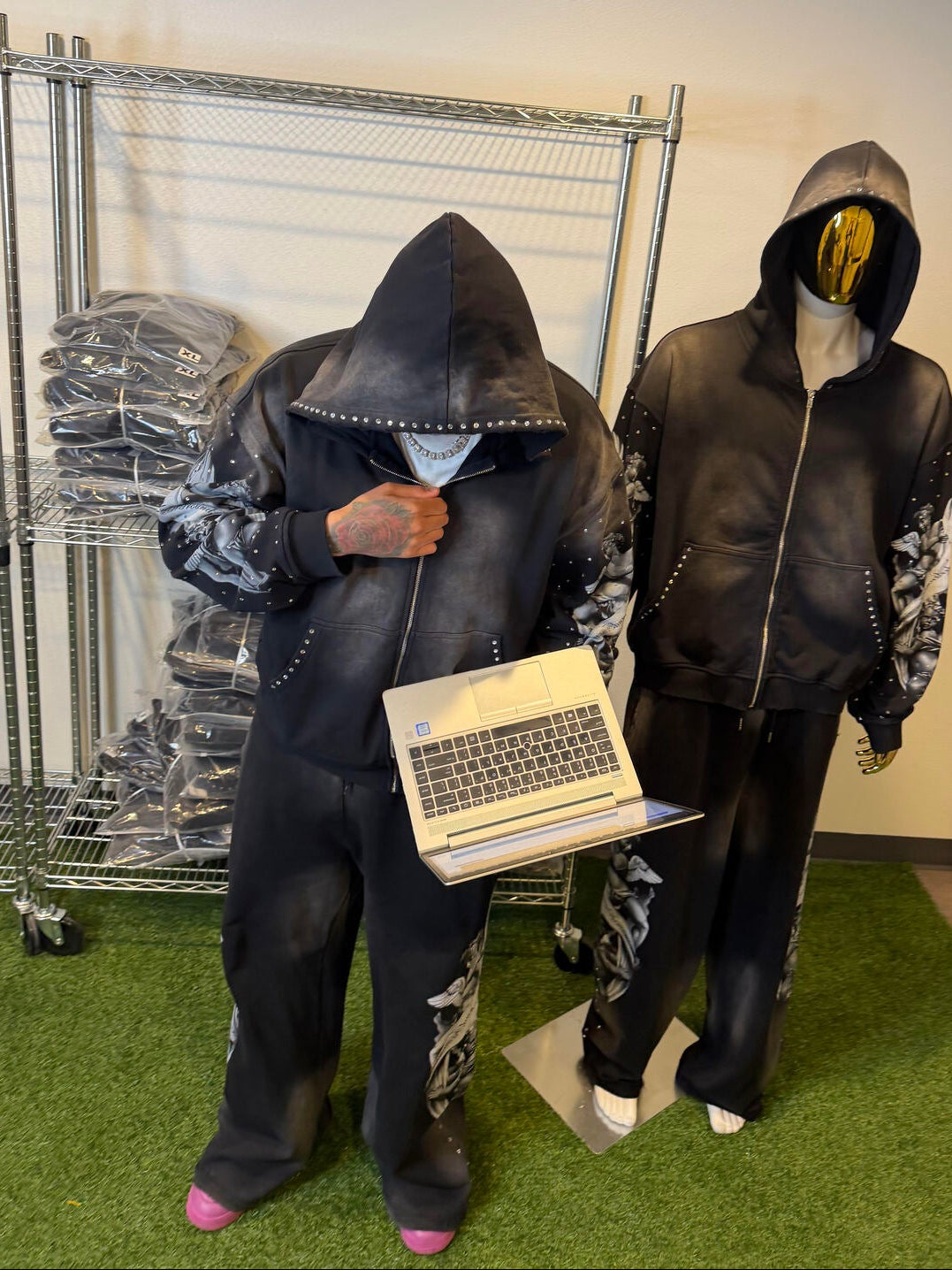 Two pwearing black hoodies with studded details, one holding a laptop, in a storage area.ople 